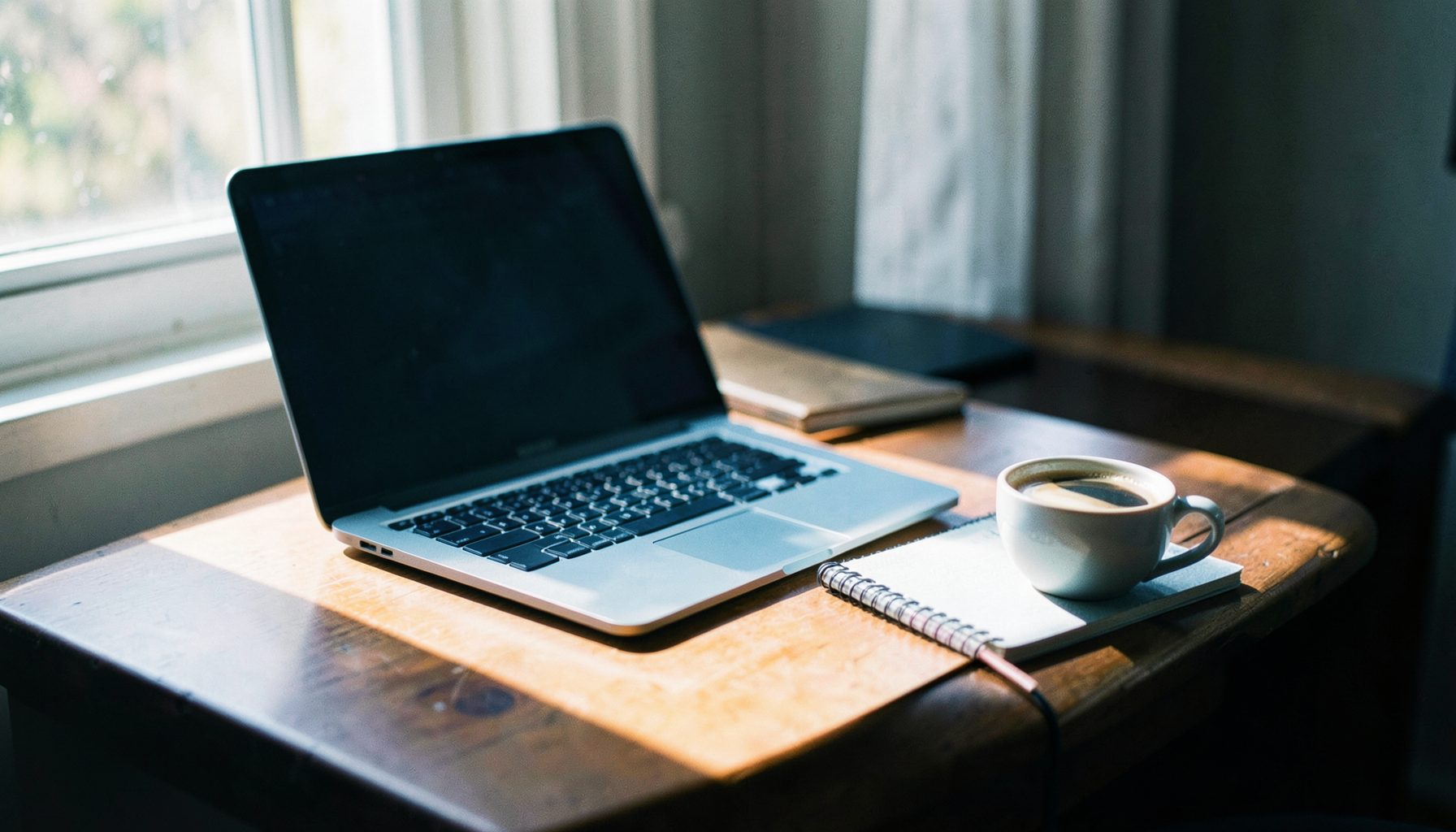A desk with a laptop, notebook, and a cup of coffee, warm morning light streaming through a window, calm productive mood