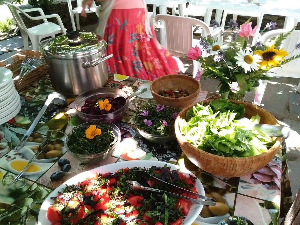 Outdoor table spread with colorful salads, herbs, edible flowers, a metal pot, and a flower bouquet, bright cheerful mood