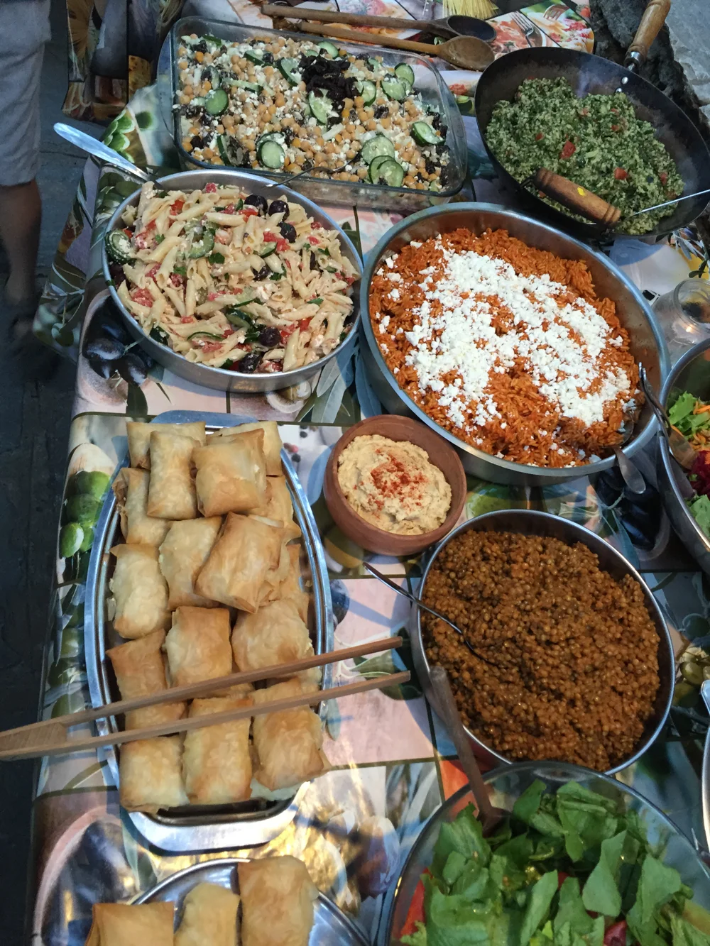 Overhead view of a table spread with various dishes including pasta salad, rice, spring rolls, hummus and lentils, warm earthy tones, casual communal dining mood