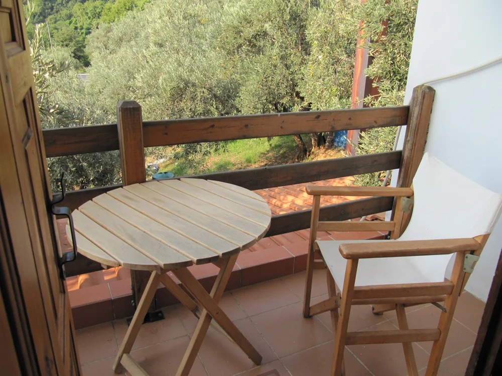 Small wooden balcony with a round table and two chairs overlooking olive trees and a tiled rooftop, warm neutral tones